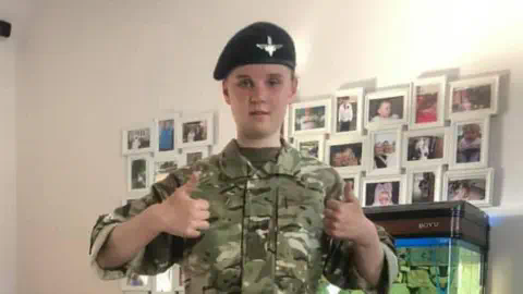 Family handout Holly Newton wearing army camouflage and a beret gives a double thumbs up to the camera. Behind her is a white wall with lots of pictures on it.